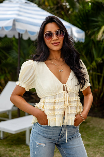 Woman wearing a white ruffled top and blue jeans standing outdoors with a striped umbrella in the background.
