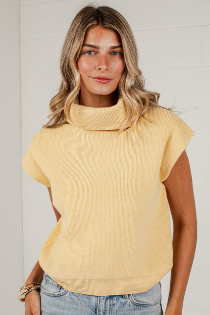 A woman wearing a pale yellow ribbed knit mock neck top with cap sleeves and light blue jeans, standing against a white brick wall.