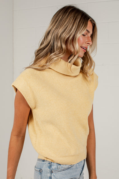 A woman wearing a pale yellow ribbed knit mock neck sweater with cap sleeves, paired with light blue jeans, standing against a white brick wall.