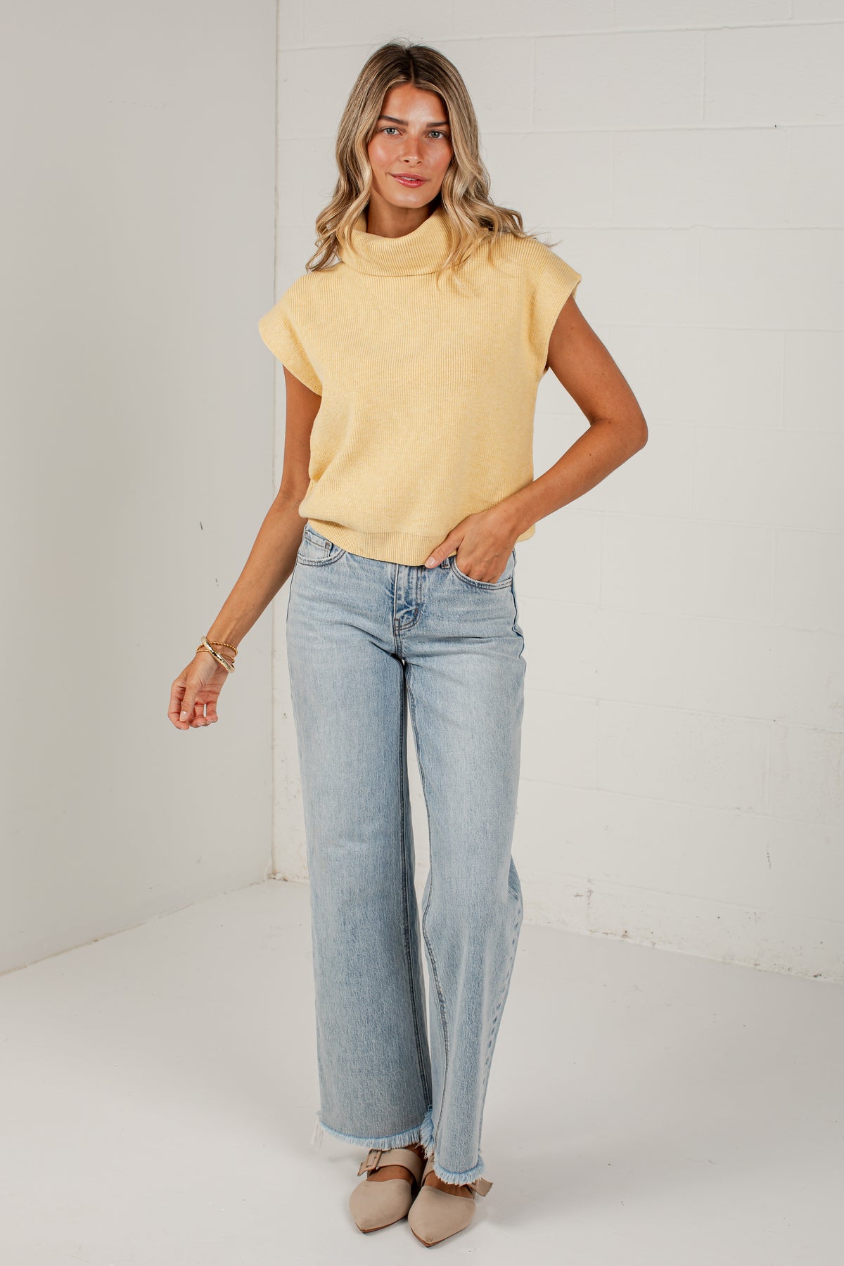 A woman modeling a pale yellow knit mock-neck top with short sleeves and light-wash wide-leg jeans, standing against a white brick wall.