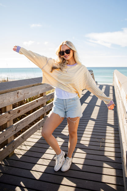 Woman standing on a wooden boardwalk by the beach, wearing a striped sweater and denim shorts.