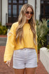 Woman wearing a yellow sweater and white shorts standing outdoors.