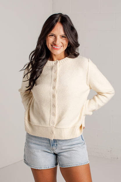 A woman wearing a cream-colored knit cardigan sweater with a button-down front and light-wash denim shorts, standing against a white wall.