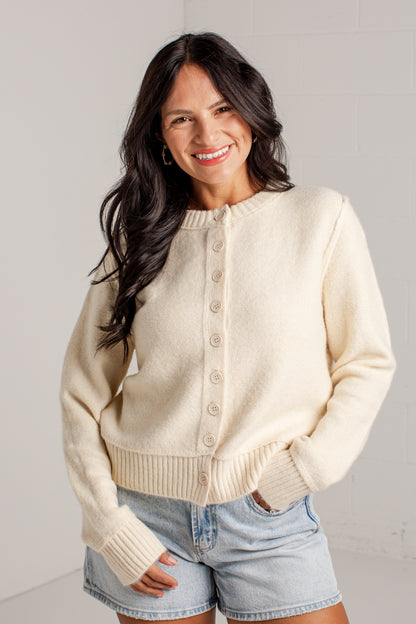 A woman wearing a cream-colored knit button-up cardigan sweater and light-wash denim shorts, standing against a light gray brick wall.