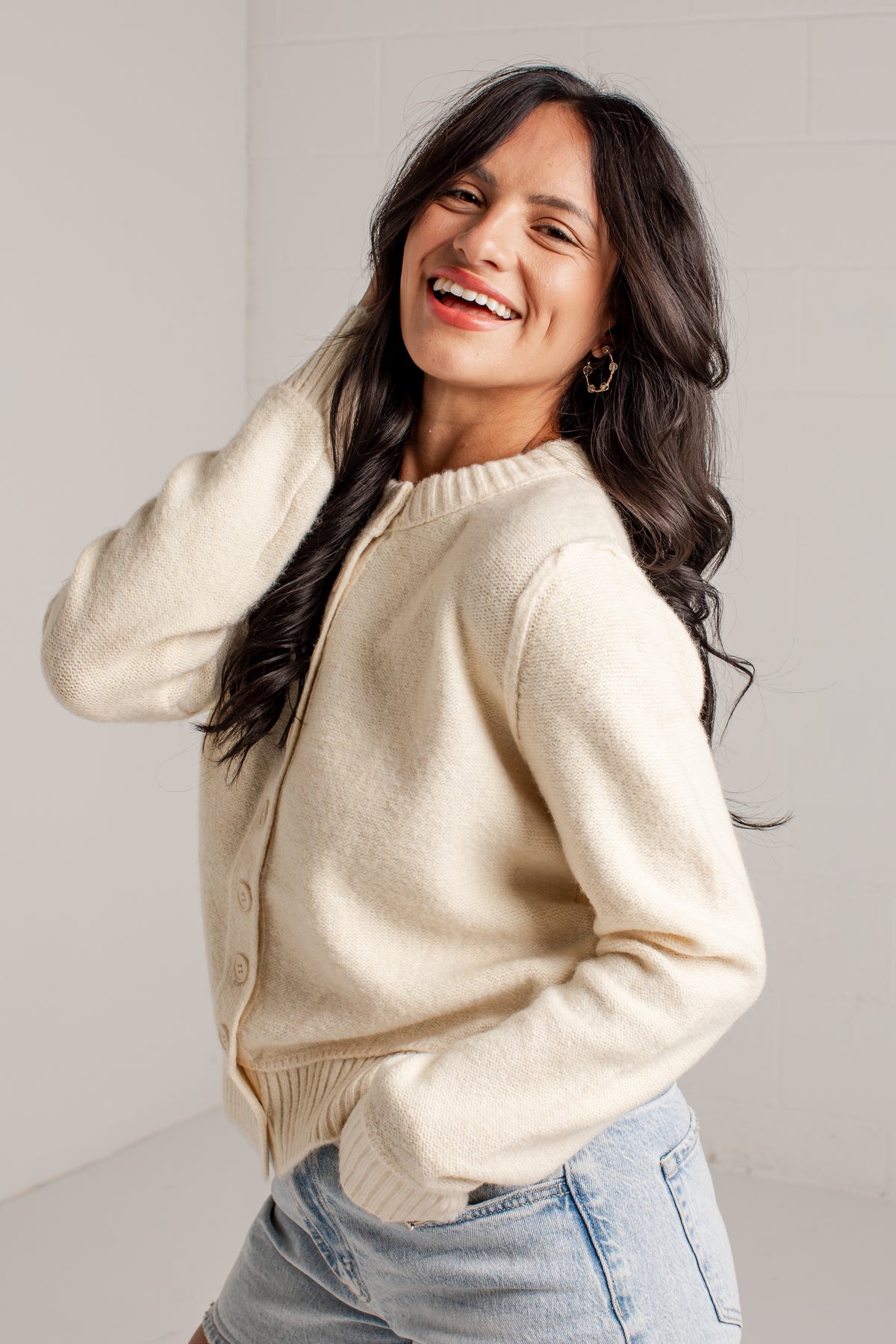 A woman smiling and looking to the side while wearing a cream-colored knit cardigan sweater and light blue denim shorts, standing against a light gray tiled wall.