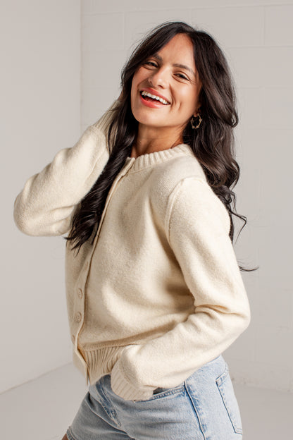 A woman smiling and looking to the side while wearing a cream-colored knit cardigan sweater and light blue denim shorts, standing against a light gray tiled wall.
