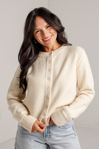 A smiling woman with long dark hair wears a cream-colored button-up cardigan sweater and light-wash blue jeans, standing against a light gray tiled wall.