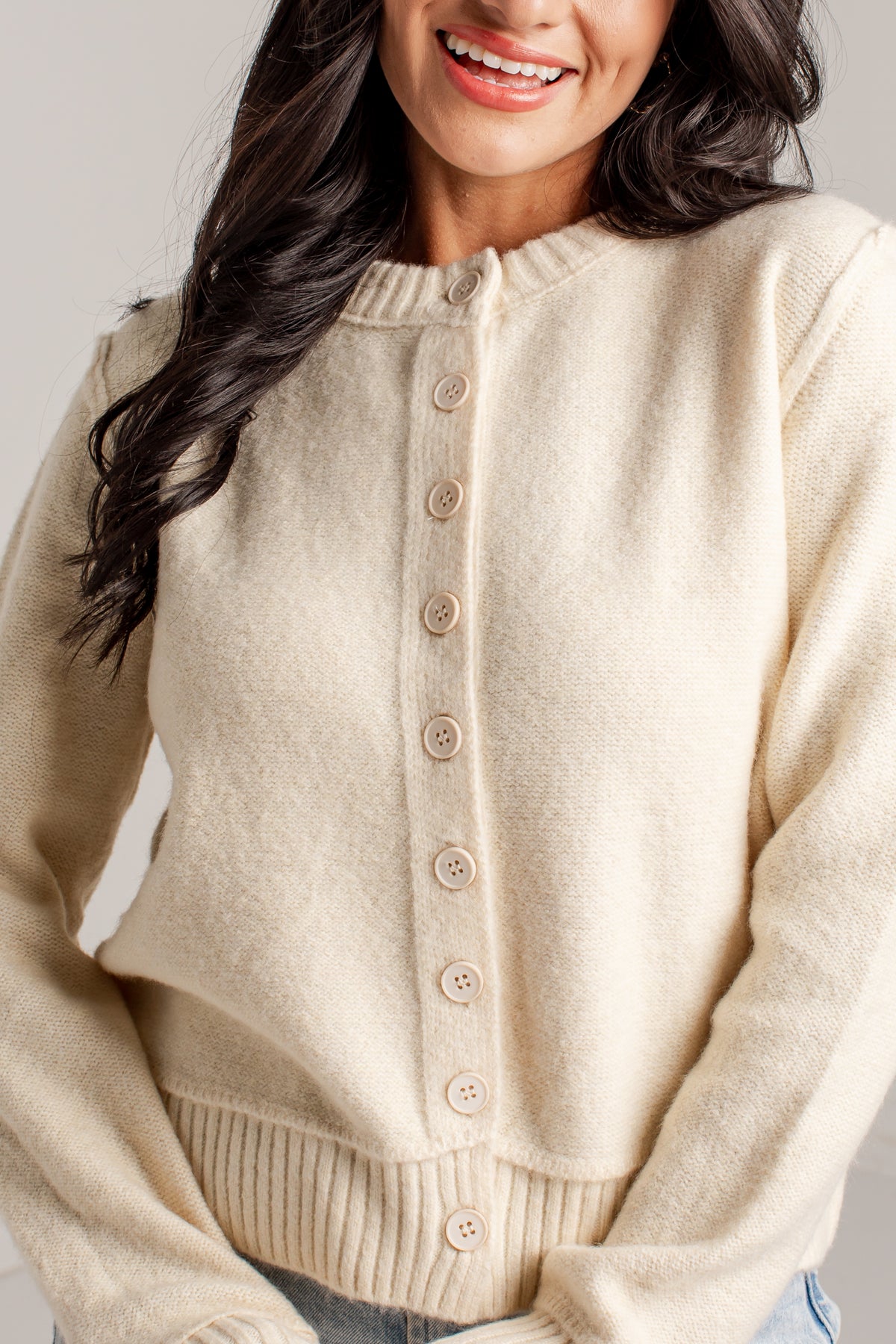 A close-up of a woman wearing a cream-colored knit cardigan sweater with a button-down front and ribbed collar, cuffs, and hem.