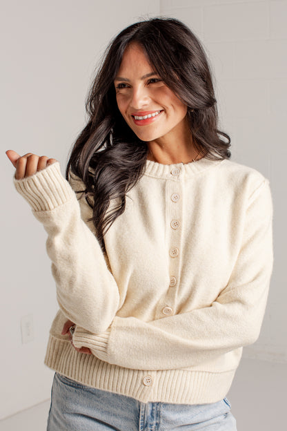 A woman wearing a cream-colored knit cardigan sweater with button-front closure and ribbed cuffs and hem, paired with light blue jeans, posing against a light gray wall.